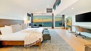 A modern bedroom with a large bed, a seating area with a view of the sea, and a TV mounted on the wall.