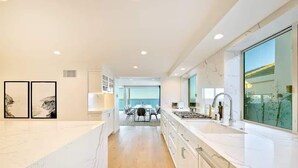 A modern kitchen with white marble countertops, a dining area with a table and chairs, and a view of the sea through large windows.