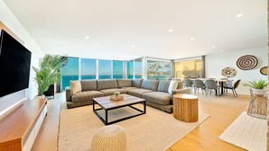 A modern living room with a large sofa, a coffee table, and a TV. There are large windows offering a view of the sea.