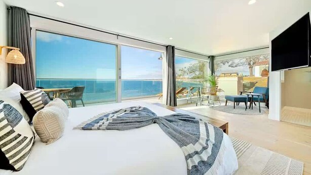 A spacious bedroom with a large bed, a balcony with a view of the sea, and a dining area with chairs and a table.
