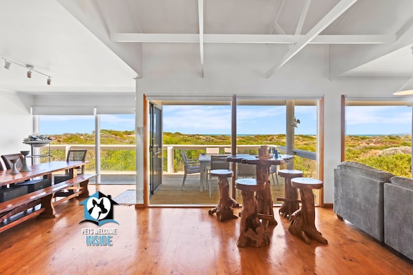 A spacious room with a wooden floor, a dining table, and a sofa. There is a large window with a view of the beach.