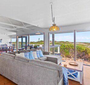 A spacious living area with a grey sofa, blue cushions, and a hanging light fixture. Large windows offer views of the outdoors.