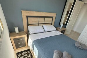 A bedroom with a wooden headboard, two white pillows, a blue bedspread, a bedside table with a lamp, and a patterned rug. - Sunset View Golf Course Apartment Alicante (Rojales)