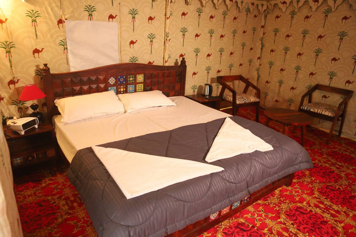 Una cama con colcha oscura y almohadas blancas, cabecera de madera con un panel decorativo, una lámpara roja, una mesita de noche con un teléfono y una alfombra estampada.