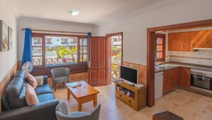 A living room with a sofa, chairs, a table, and a TV. There is a kitchen with wooden cabinets and a stove. - Casa del Mar Ilic - Communal pool, Sea Views, 10 Minutes to Marina Rubicón (Playa Blanca)