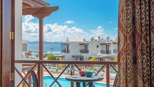 A balcony with a pool, a view of the sea, and buildings in the background. - Casa del Mar Ilic - Communal pool, Sea Views, 10 Minutes to Marina Rubicón (Playa Blanca)