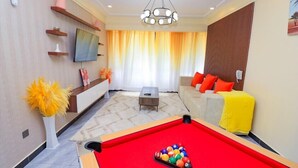 A modern living room with a pool table, a sofa with orange cushions, a TV mounted on the wall, and a decorative plant. - Staroot Residences (Nairobi)