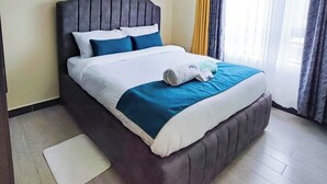 A neatly made bed with a headboard, a white pillow, a blue pillow, and a teal blanket. - Staroot Residences (Nairobi)