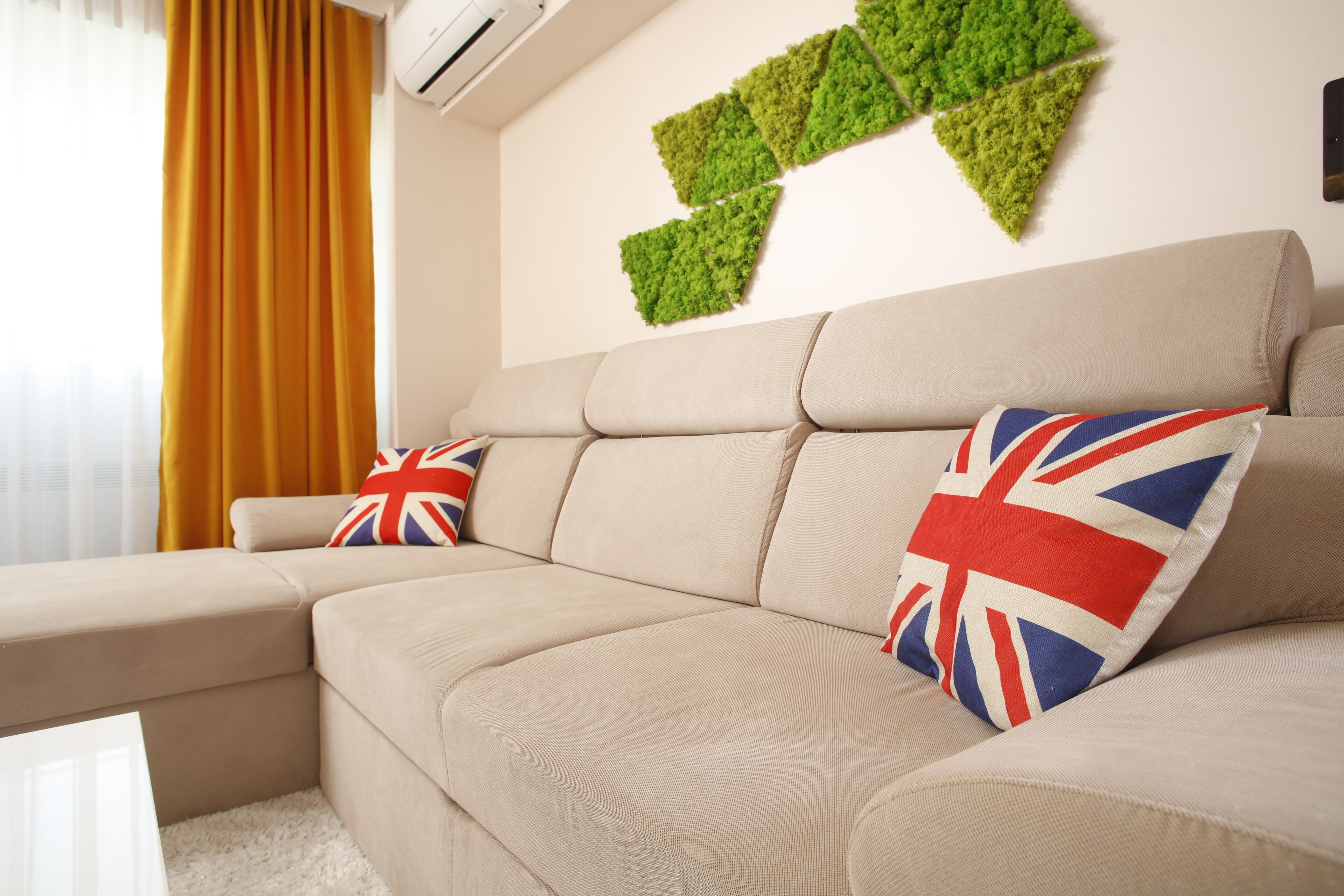 A beige sectional sofa with British flags on pillows, a green moss wall art, and a window with yellow curtains.