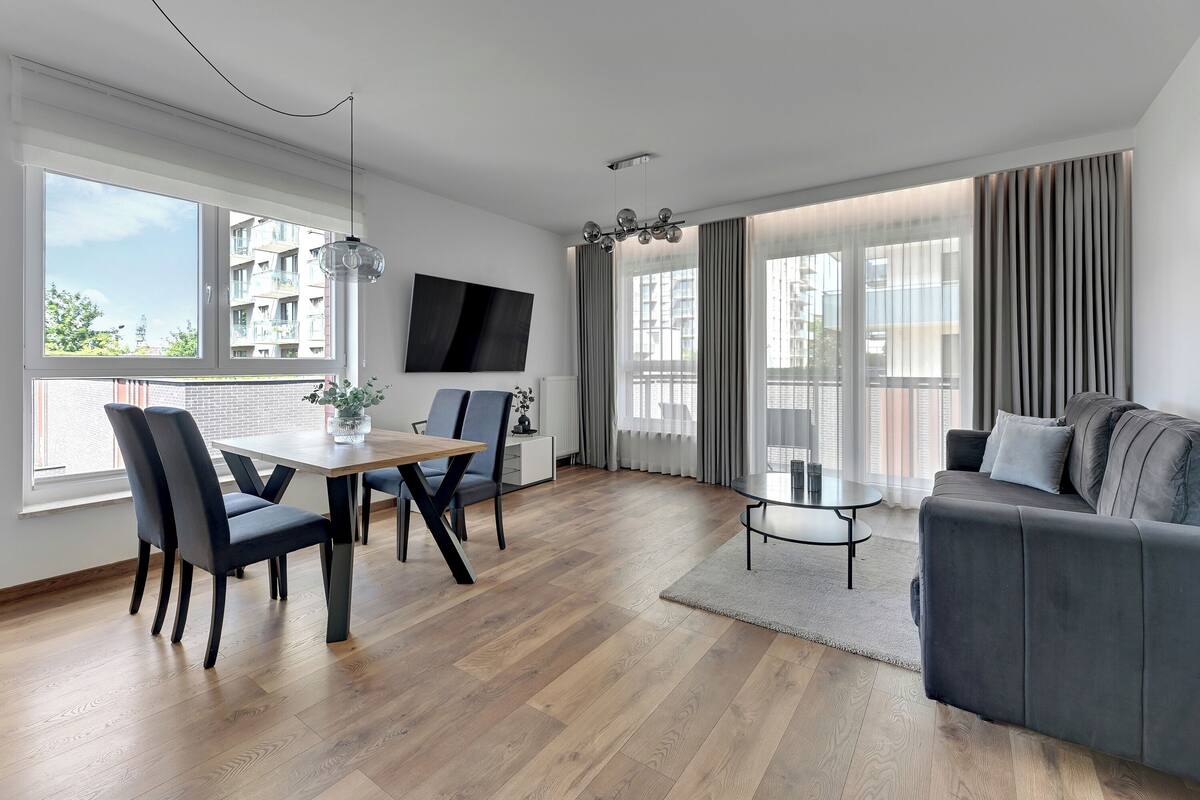 A modern living room with a dining area, a grey sofa, and a flat-screen TV mounted on the wall.