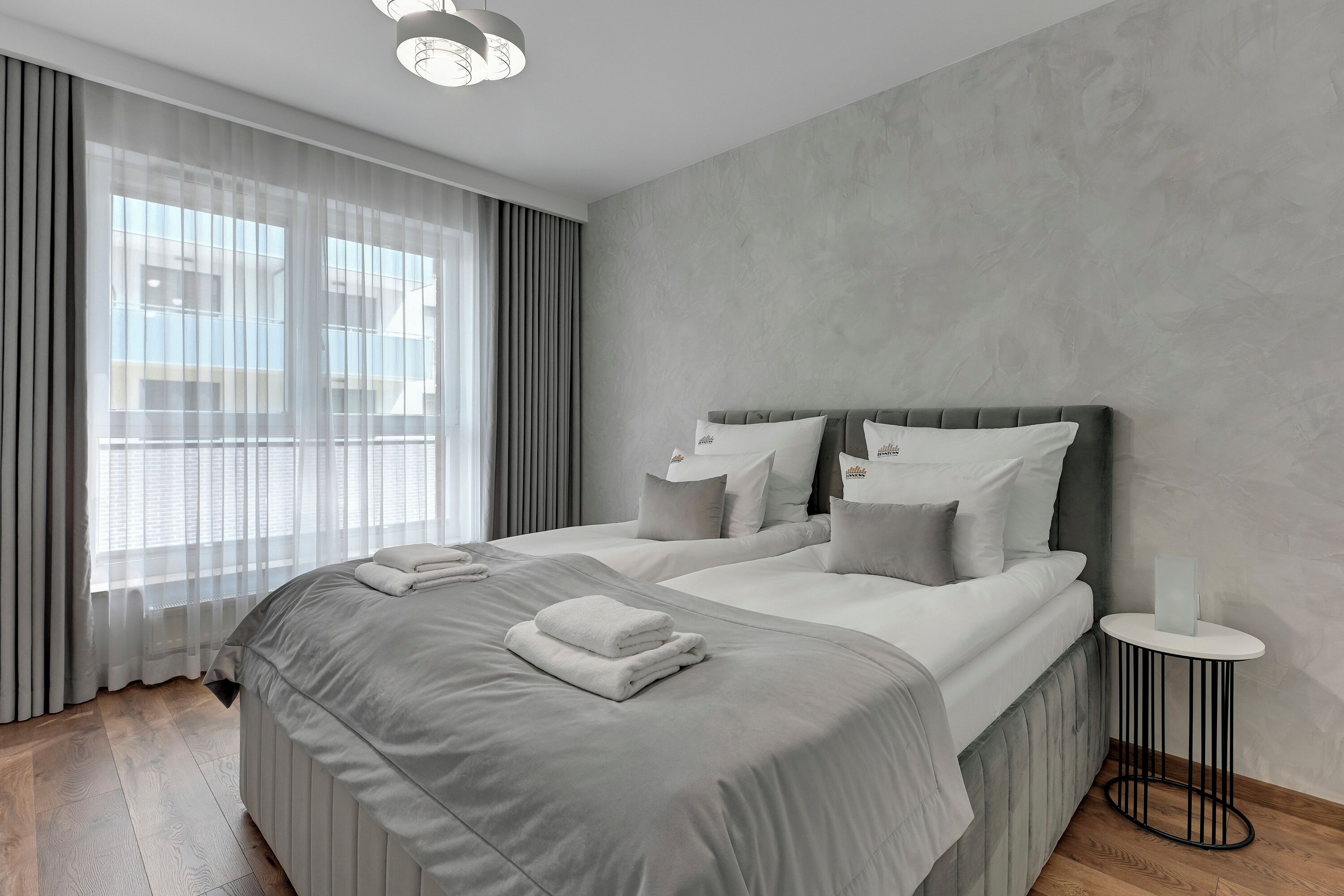 Une chambre d’hôtel moderne avec un grand lit, une literie grise, une table d’appoint ronde et des rideaux transparents.