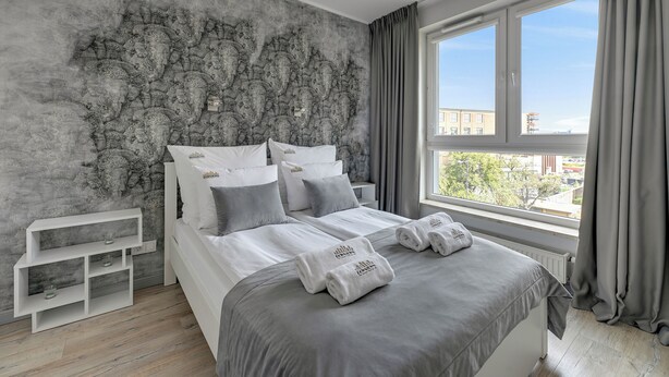 A modern bedroom with a grey and white color scheme, featuring a large bed with grey bedding, a white nightstand, and a view of the city through a large window. -  Bastion Wałowa City Center & Parking by Downtown Apartments (Gdansk)