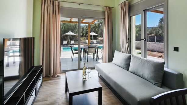 Ein modernes Wohnzimmer mit einer Couch, einem Couchtisch und einem Fernseher. Eine Glastür öffnet sich zu einem Außenbereich mit Pool und Sonnenschirmen.