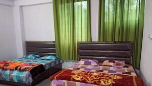 A room with two beds, each with a distinct patterned bedspread. The room has green curtains and a plain white wall. - Indralok Hotel By GRB (Jakholi)