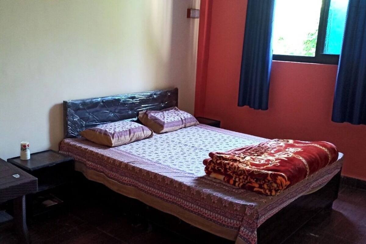 Un lit simple avec une couvre-lit à motifs et des oreillers, une petite table de chevet avec une lampe, et une fenêtre avec des rideaux.