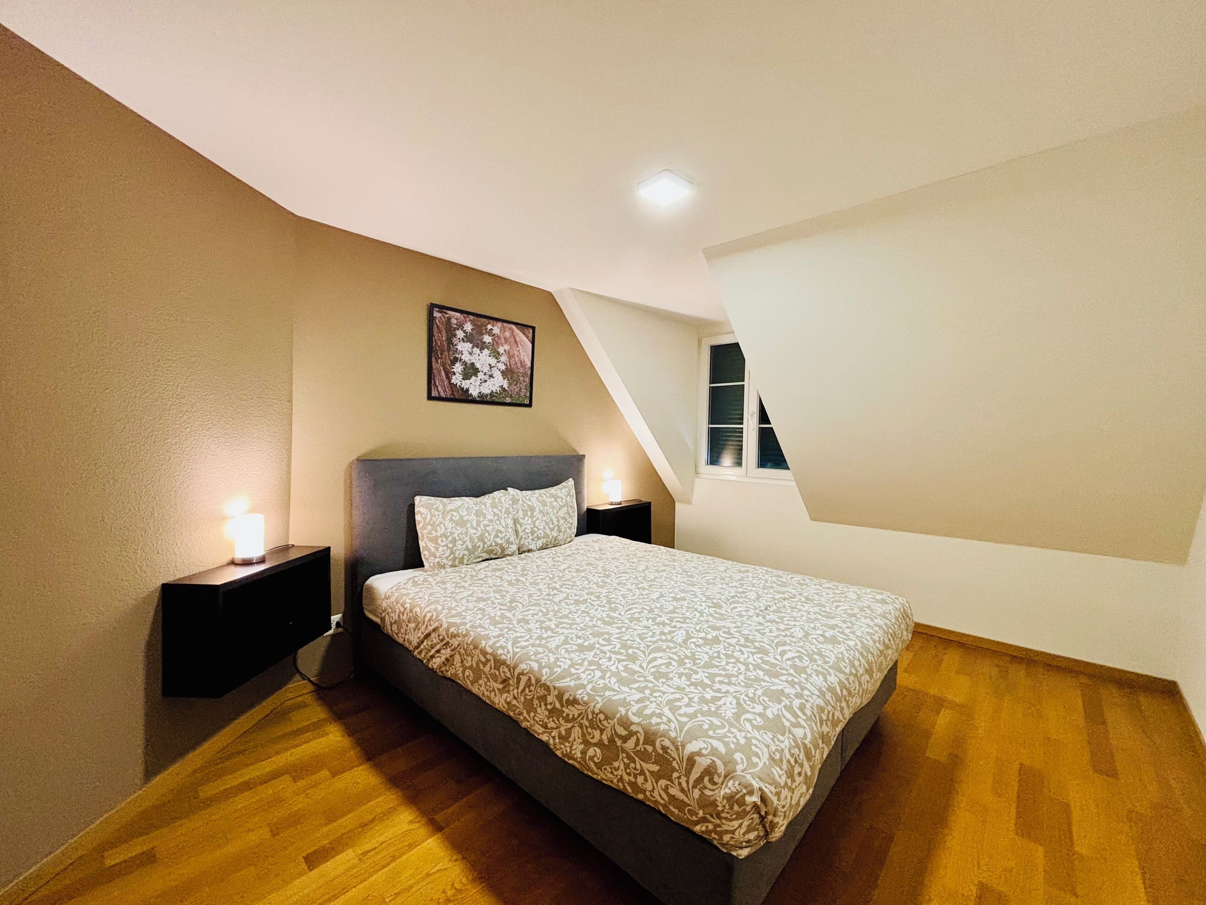 Un dormitorio con cama, mesitas de noche, un cuadro en la pared y techo inclinado.