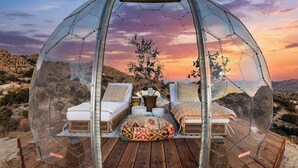 Estrutura em cúpula transparente com duas áreas de assento acolchoadas em branco, um tapete de palha trançada e uma pequena mesa com uma vela.