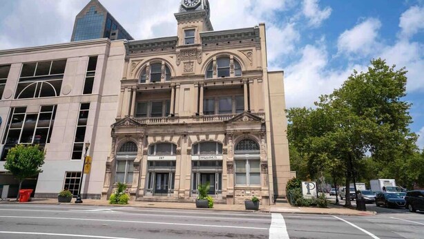 Un edificio histórico con una torre de reloj, grandes ventanales y un llamativo cartel con la dirección.