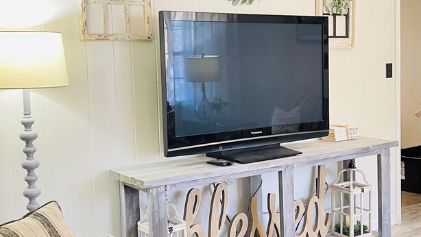 A living room with a television on a wooden console, a lamp, and decorative wall elements. - 3-Bdrm Home on Large Property at Norris Lake. Pet Friendly! (New Tazewell)