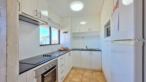 Cocina compacta con gabinetes blancos, encimera negra, horno integrado y ventana con vistas al exterior.