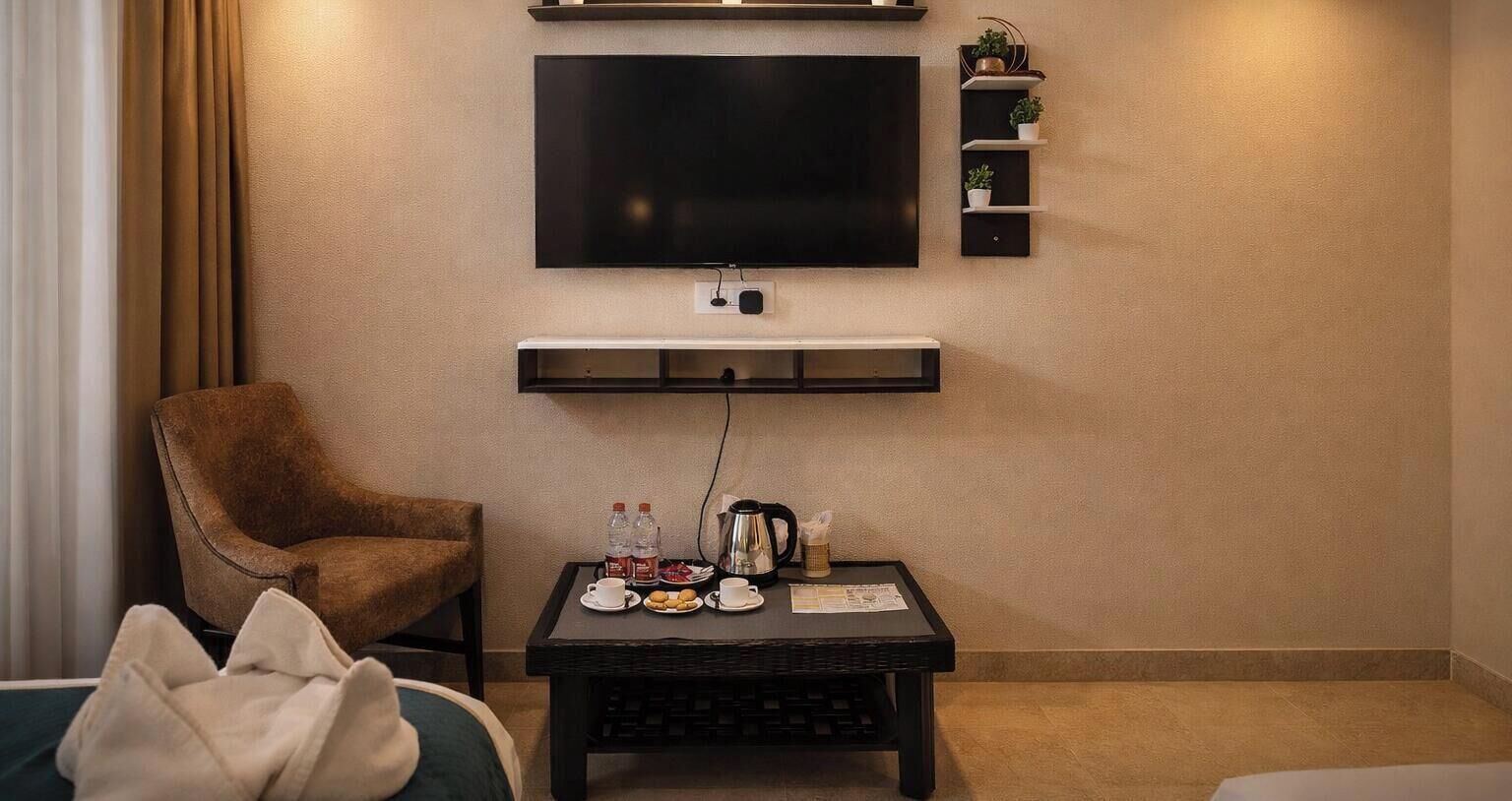 Una camera d'albergo con una TV a schermo piatto montata a parete, una poltrona marrone, un tavolino con un vassoio di snack e uno scaffale con piante in vaso.