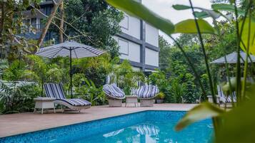 Relax by the serene pool surrounded by lush greenery and shaded by striped umbrellas.
