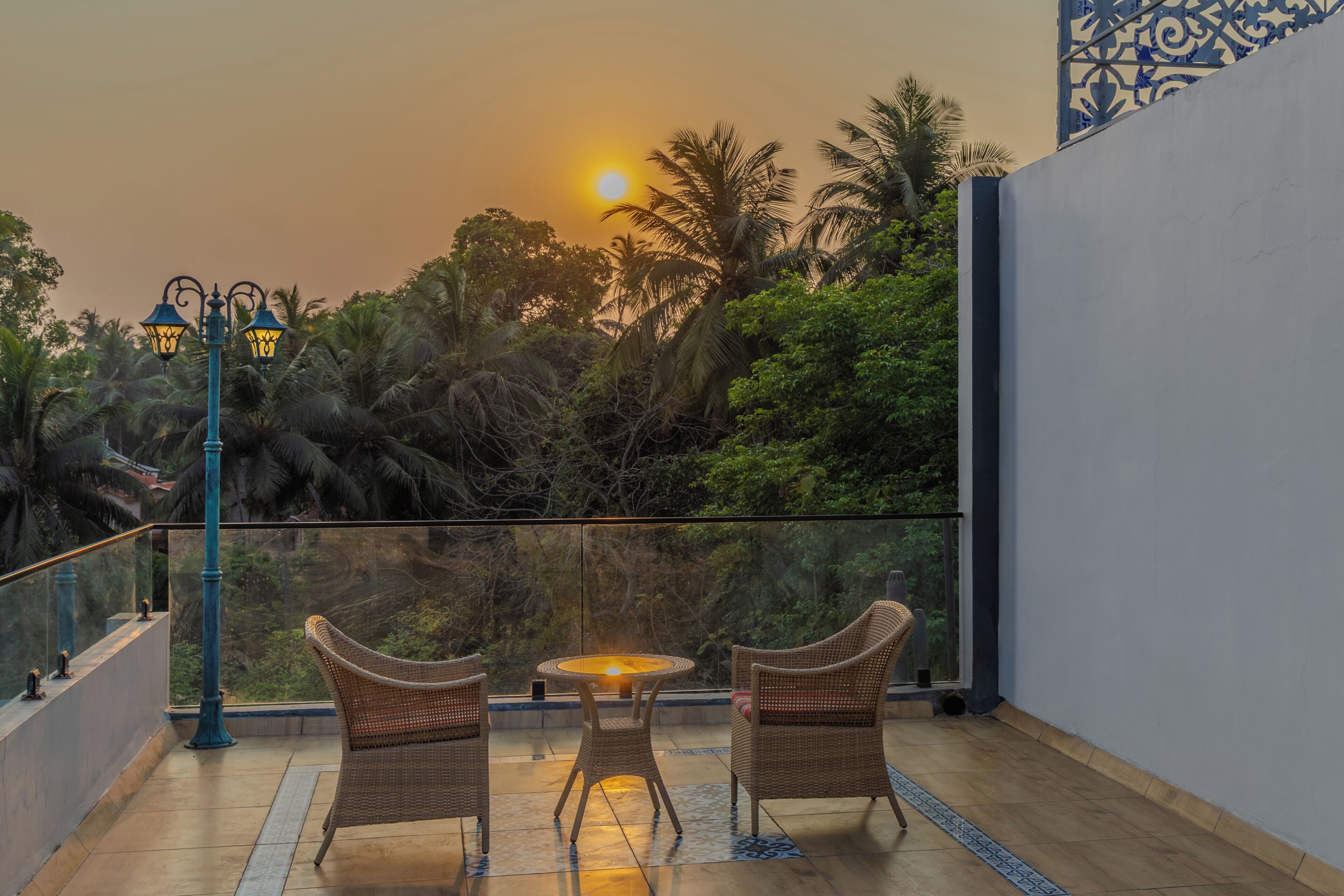 Un balcón con mobiliario de mimbre, una mesa con lámpara y vistas a palmeras y al atardecer.