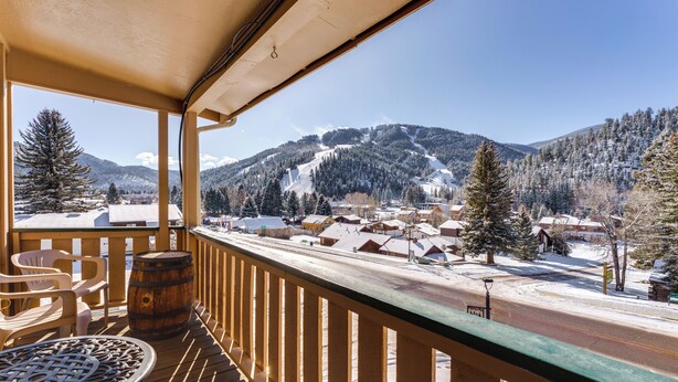 Um deck de madeira com mesa e cadeiras, com vista para uma cidade coberta de neve e montanhas ao fundo.