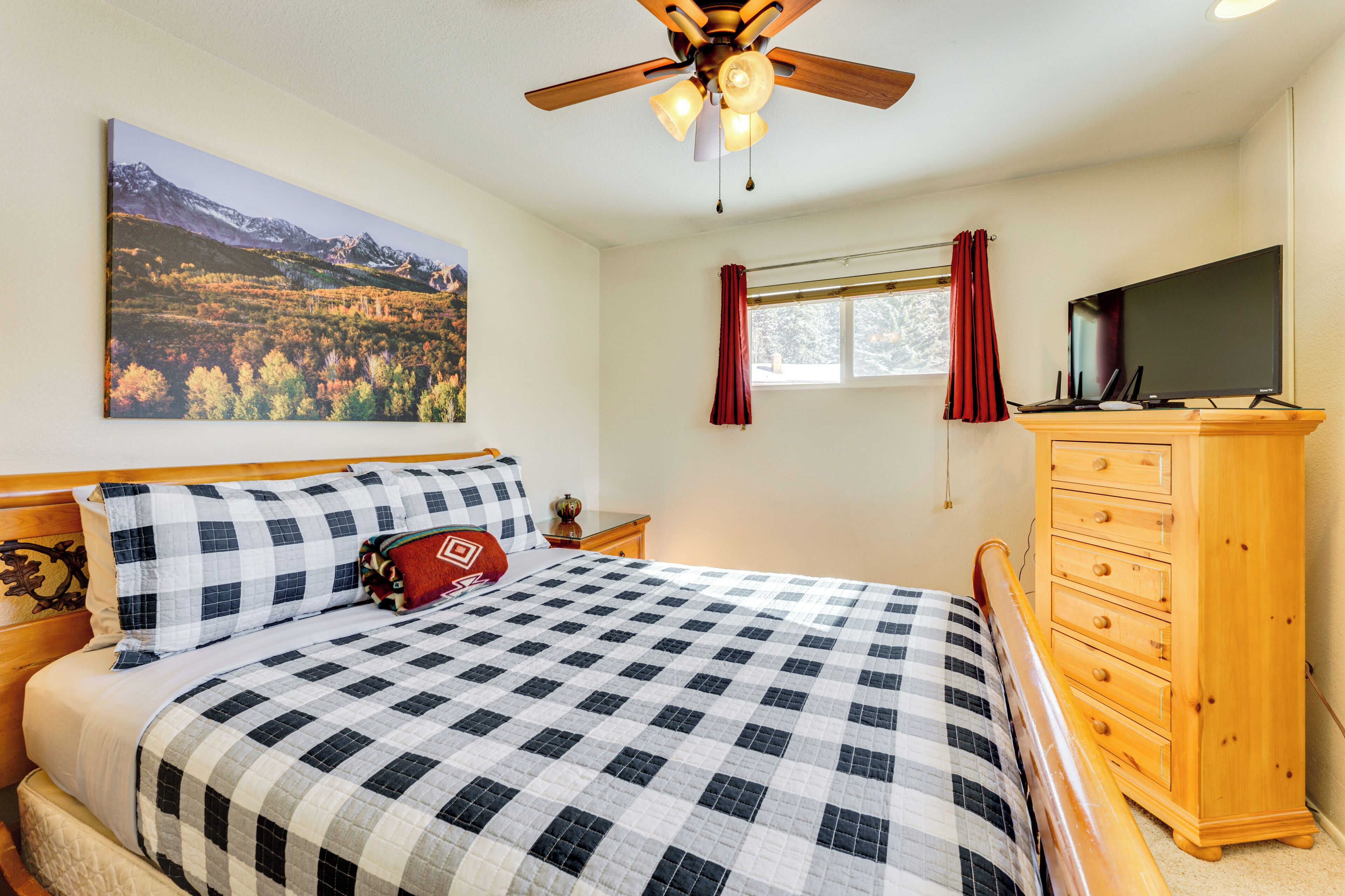 A bedroom with a checkered bedspread, a wooden dresser, a flat-screen TV, a ceiling fan, a window with red curtains, and a painting of a mountain landscape.