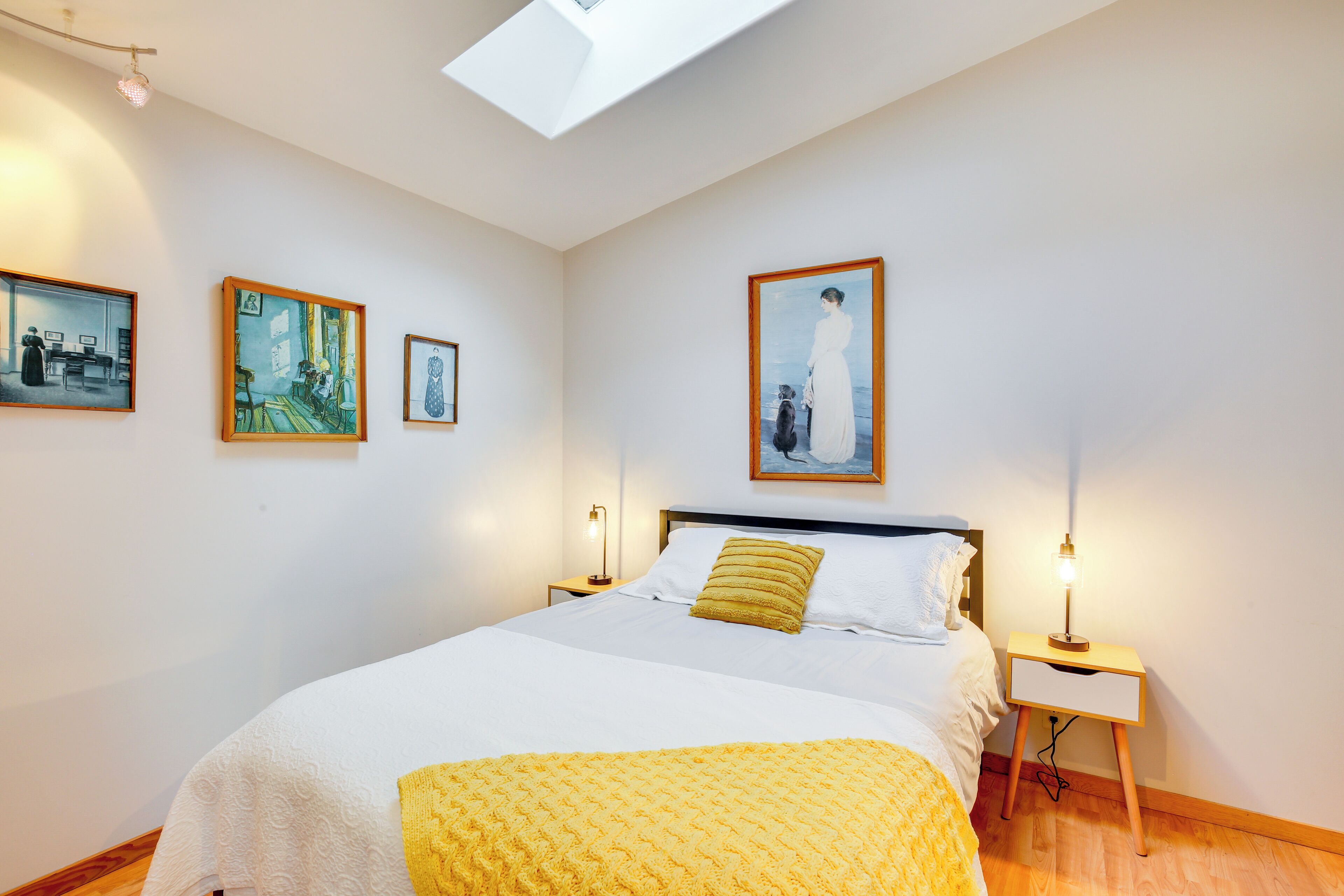 A bedroom with a bed, a bedside table with a lamp, and three framed artworks on the wall.