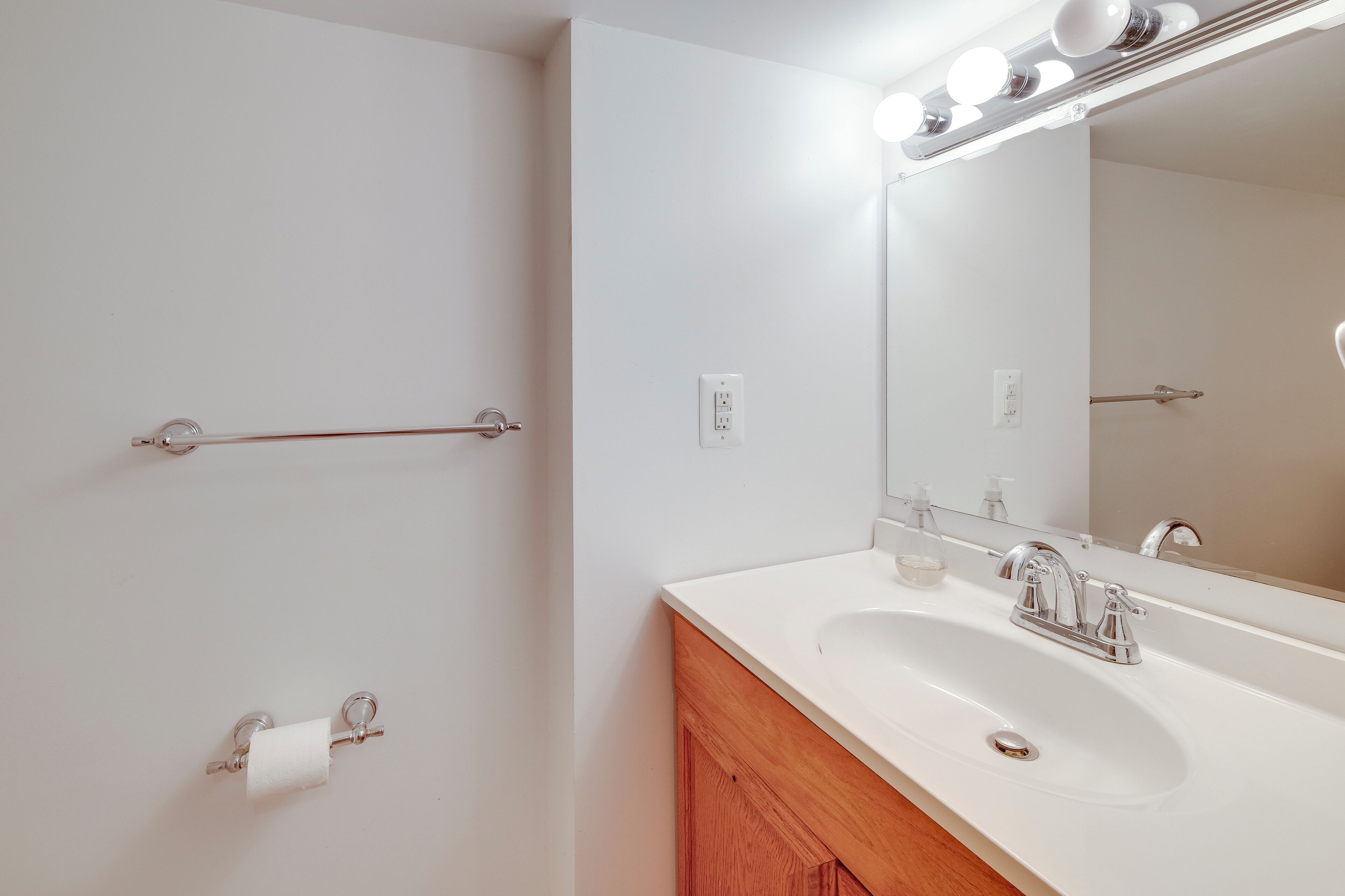 Un baño con lavamanos blanco, un espejo con luces, un perchero para toallas y un porta papel higiénico.