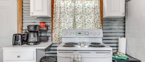 A kitchen with a stove, a coffee maker, and a trash can.