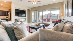 A spacious living room with a large sofa set, a coffee table, and a flat-screen TV mounted on the wall. There is a sliding glass door leading to an outdoor patio area with a dining table and chairs. - Covered Patio & Community Pool: Home in Surprise (Surprise)