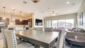 A spacious dining area with a large table and chairs, a kitchen in the background, and a view of the outdoors through large windows. - Covered Patio & Community Pool: Home in Surprise (Surprise)