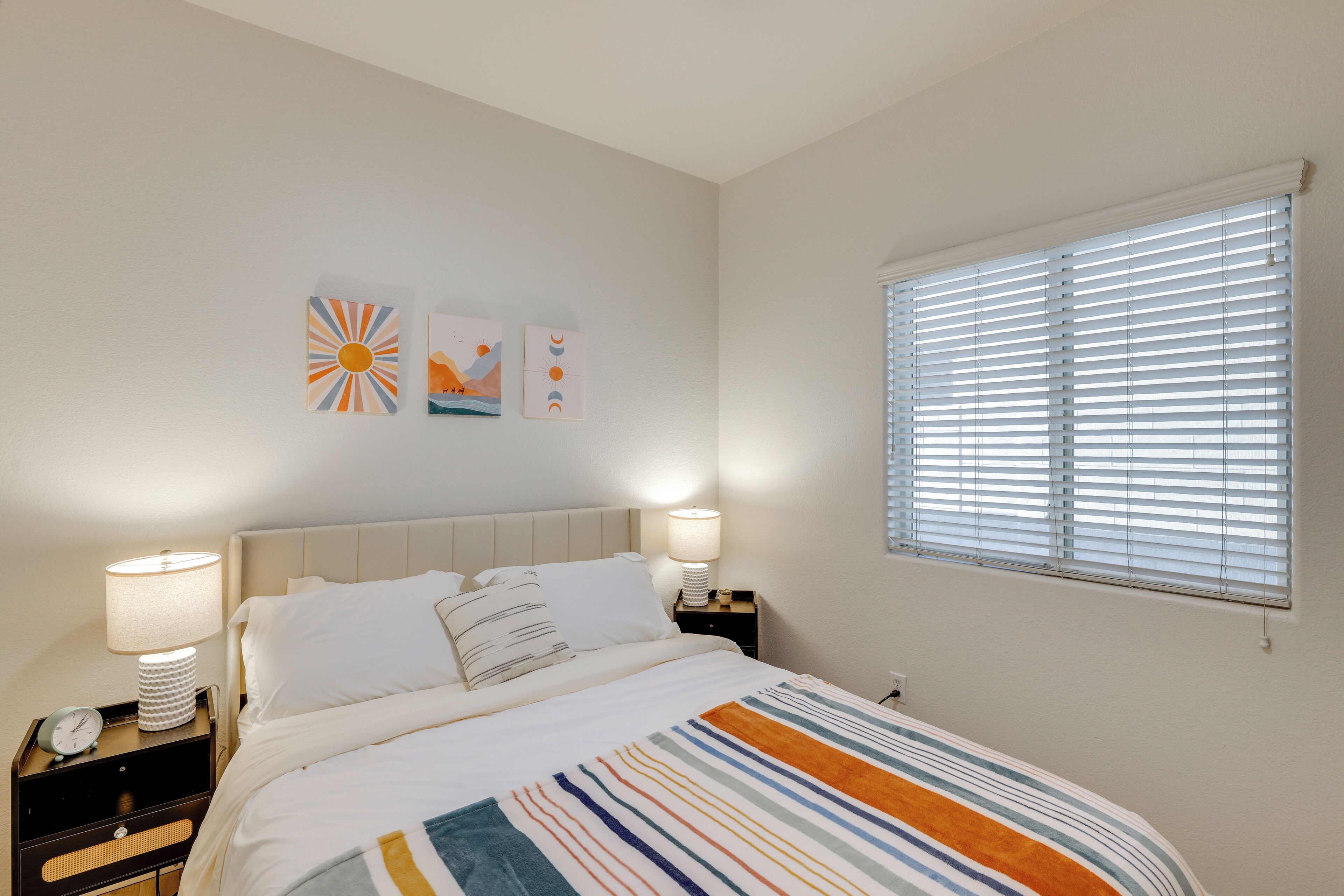 A bedroom with a bed, two bedside tables, a window with blinds, and wall art.
