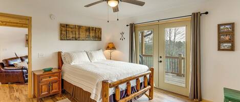 A bedroom with a wooden bed, a ceiling fan, a nightstand, a green table, and a large window with curtains.