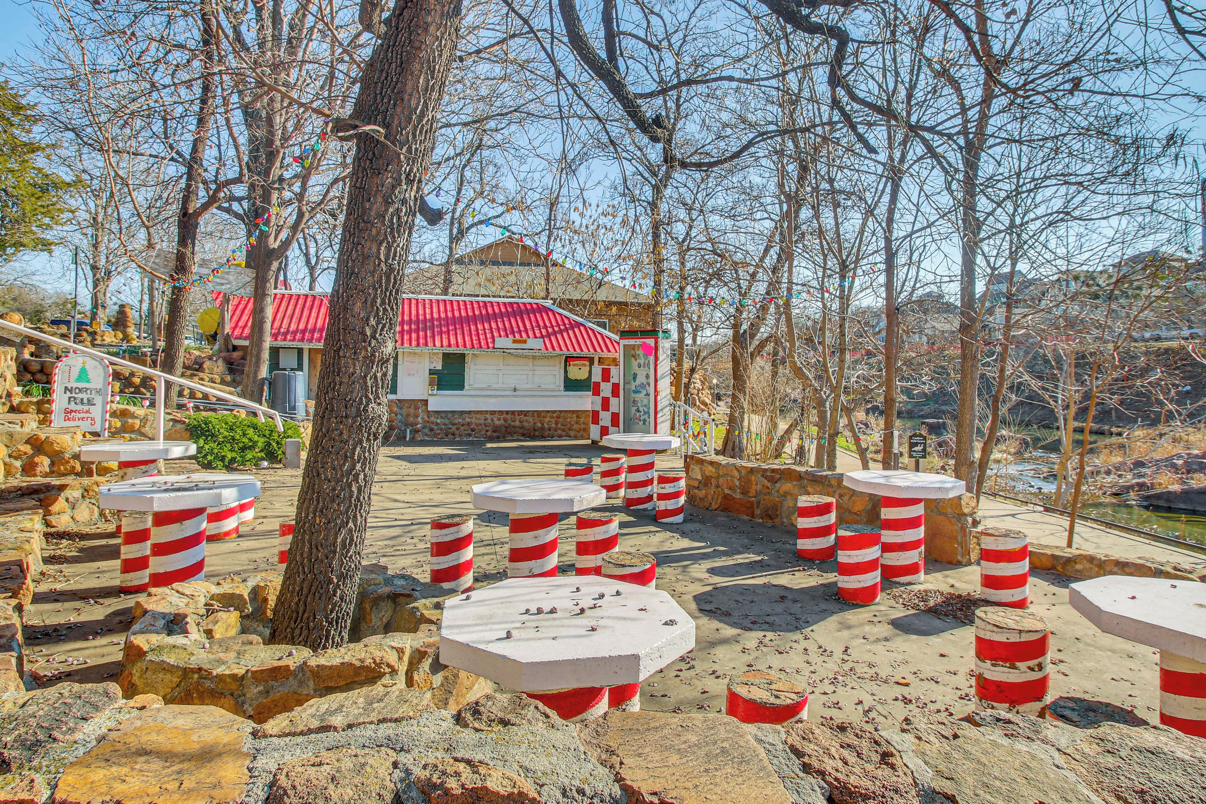 A rustic outdoor seating area with stone tables and red-striped stools, surrounded by trees and a building with a red roof.