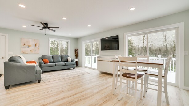 A spacious living room with a grey sofa, a dining table with chairs, a ceiling fan, and large windows offering a view of trees. - 3 Mi to Lake Huron: Secluded Home in Rogers City (Rogers City)