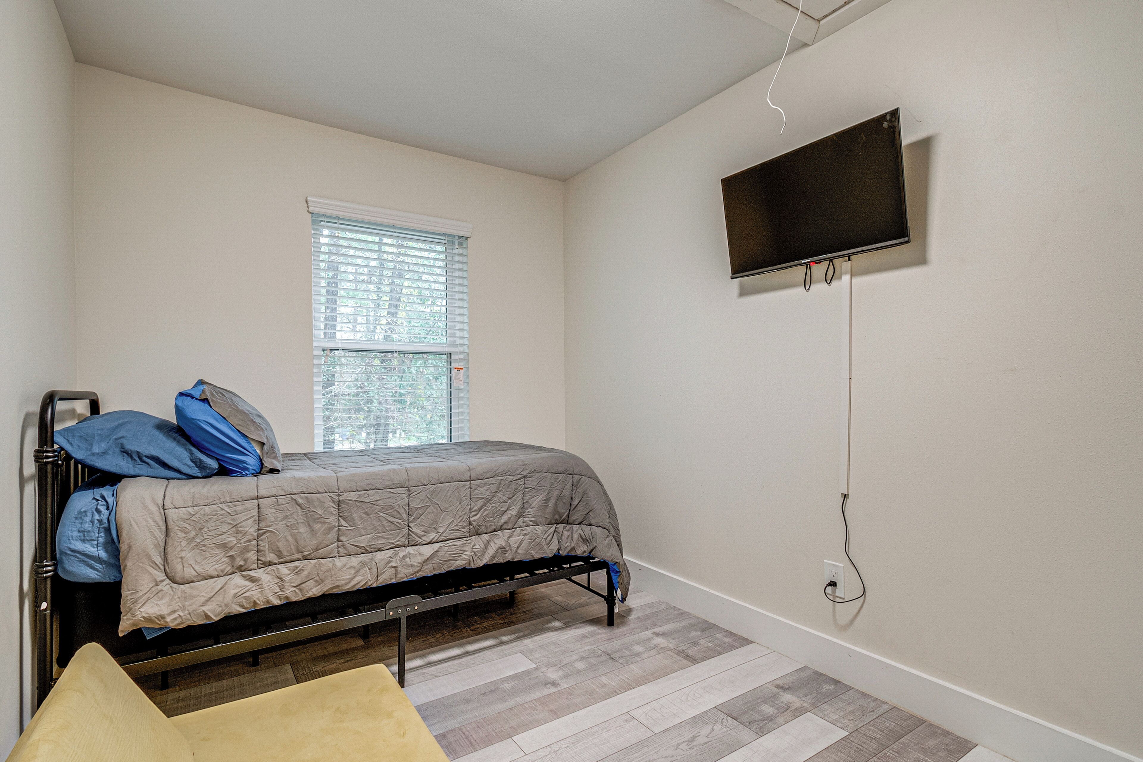 Ein Schlafzimmer mit einem Bett, einem Fenster mit Jalousien, einem an der Wand montierten Fernseher und Holzboden.