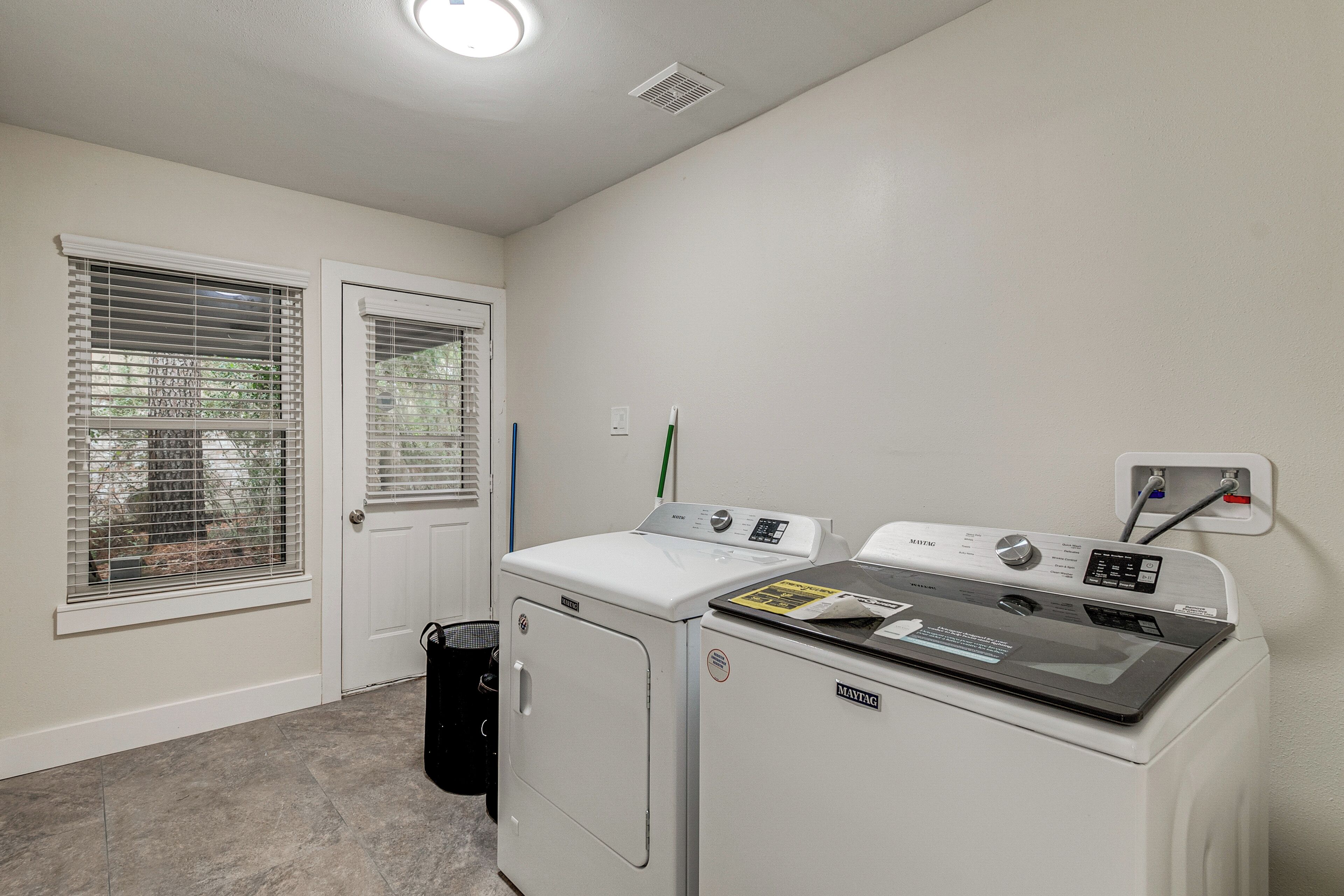 A laundry room with a front-loading washer and dryer, a trash can, and a window with blinds.