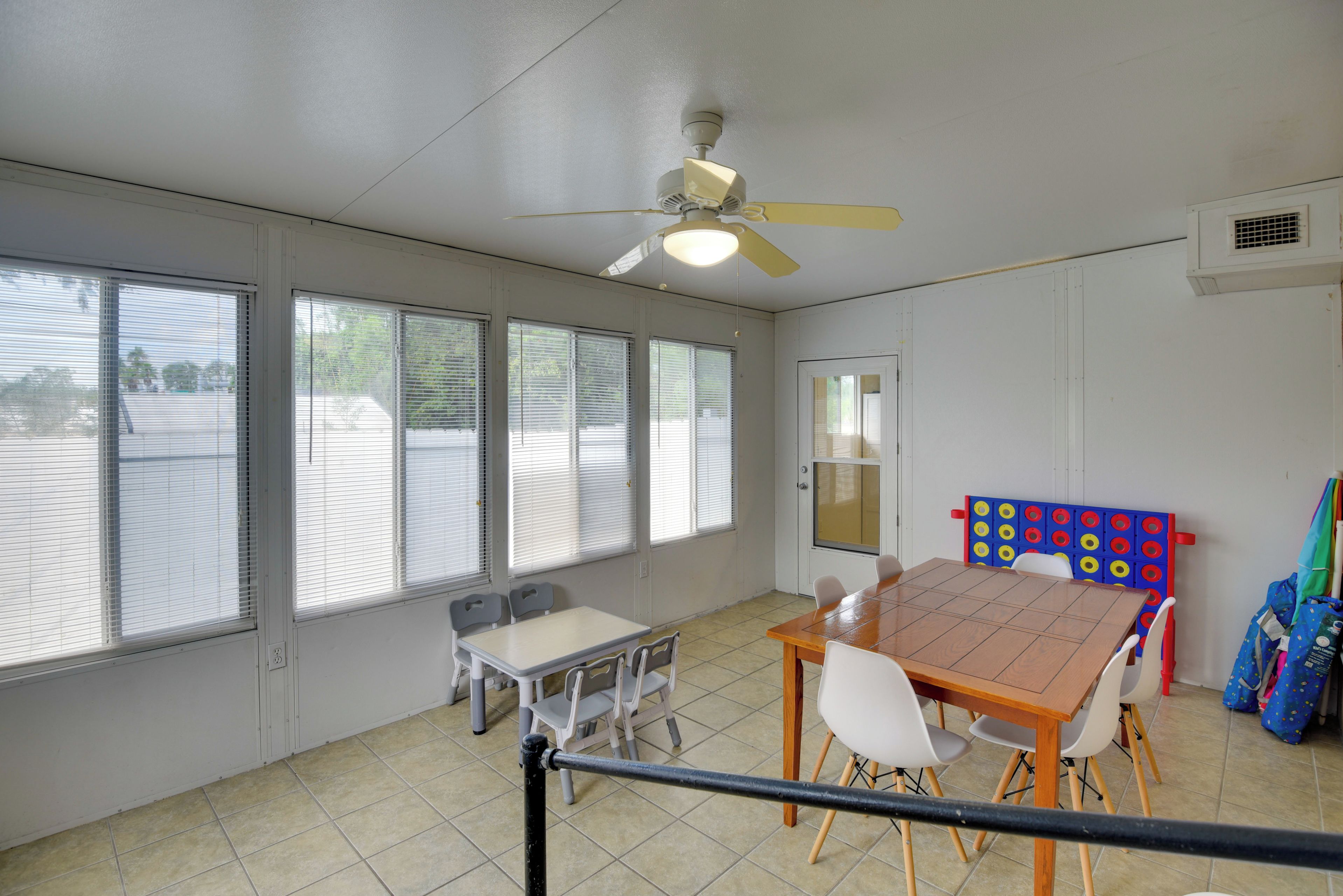 Una habitación con una mesa y sillas, ventanas amplias, un ventilador de techo y una puerta con ventanal.
