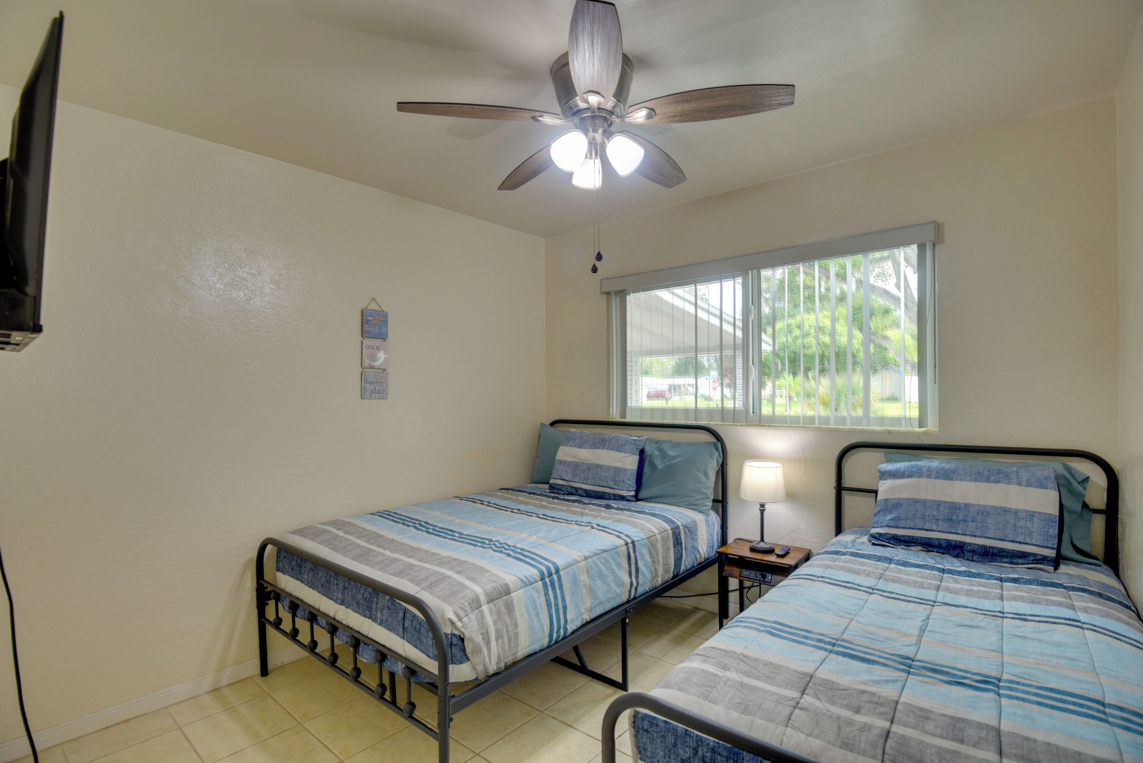A bedroom with two beds, a ceiling fan, a window with blinds, and a wall-mounted TV.
