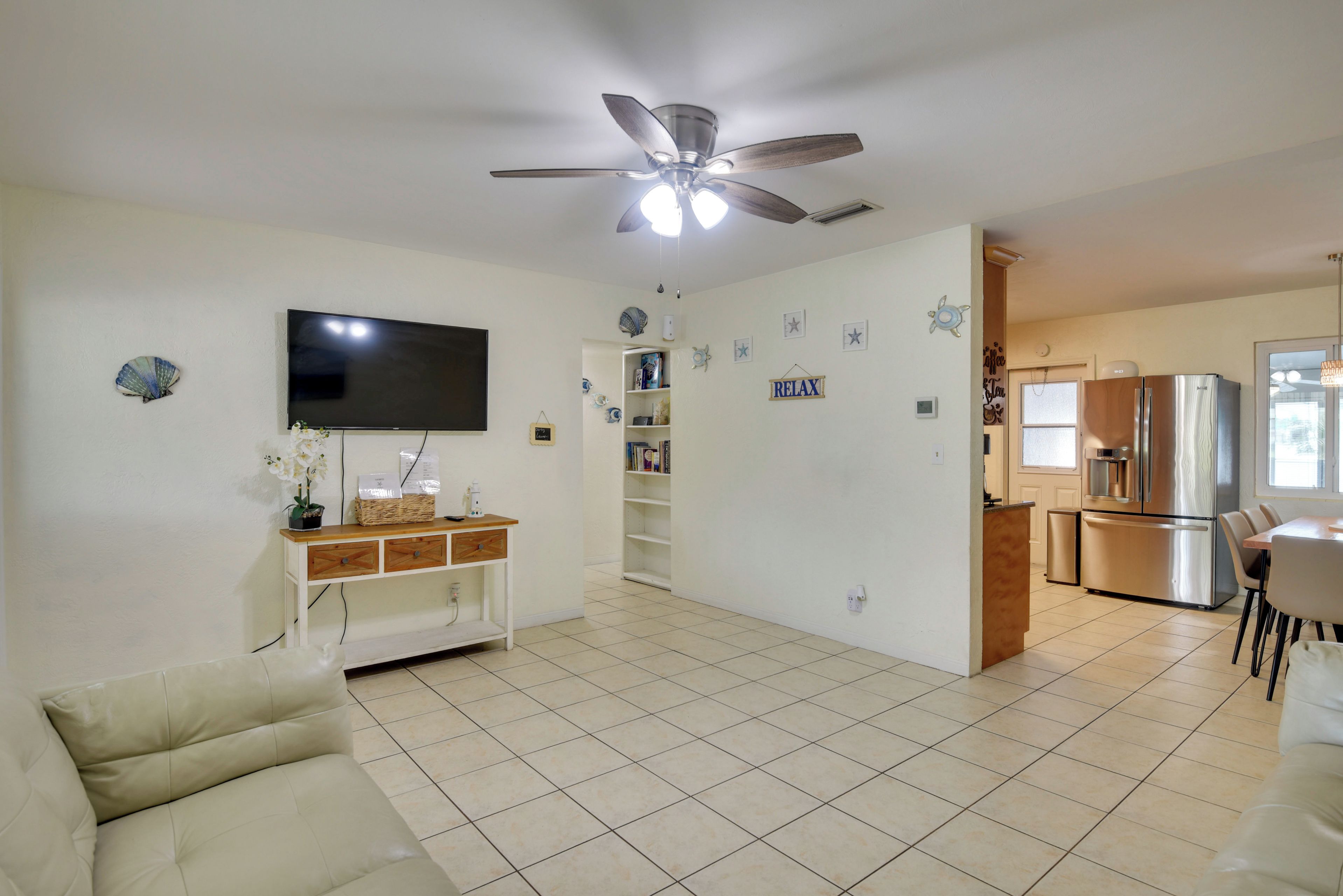 A living room with a flat-screen TV, a ceiling fan, a sofa, and a kitchen area with a refrigerator and a microwave.