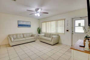 A living room with two beige sofas, a ceiling fan, a window with blinds, and a door with a decorative item. - 4 Mi to Shore: Family Home in Daytona Beach (Holly Hill)
