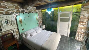 Un dormitorio con pared de piedra, una cama con ropa blanca, cabecero de madera y una puerta que da a un baño.