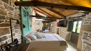Una habitación rústica con pared de piedra, vigas de madera en el techo, una cama con ropa blanca, una máquina de coser antigua y un televisor sobre una pared de piedra.