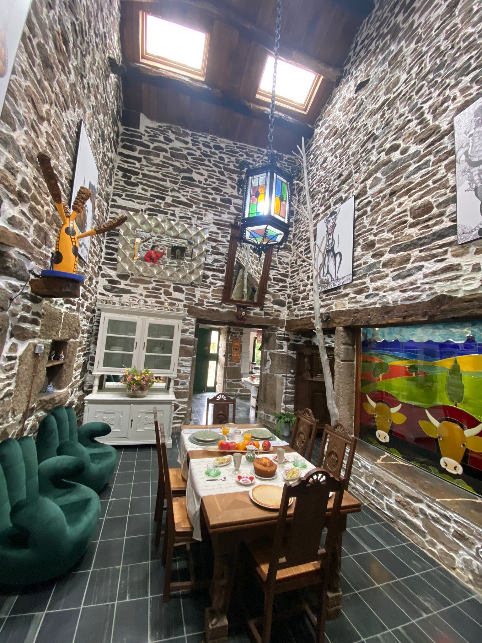 A rustic stone-walled room with a dining table set for breakfast, a green sofa, and a colorful mural on the wall.