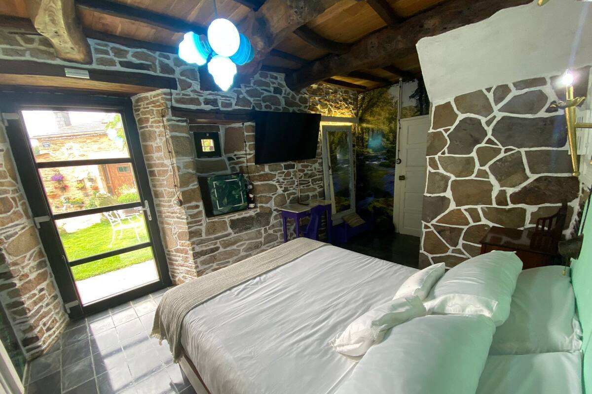 A bedroom with a stone wall, a bed with white linens, a television mounted on the wall, and a window with a view of the garden.