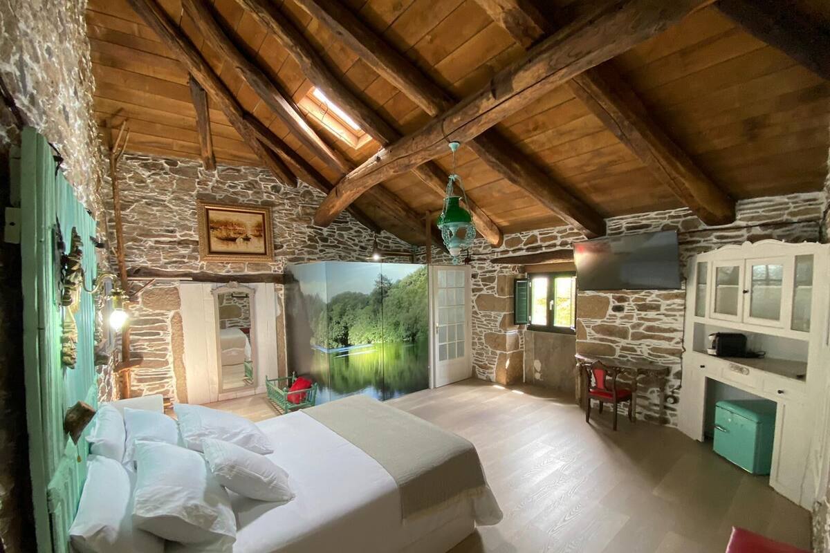 A rustic bedroom with a stone wall, wooden beams, a bed with white linens, a green landscape mural, a small table, and a cabinet.
