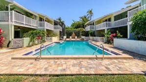Une piscine entourée de bâtiments à deux étages dotés de balcons, sous un ciel dégagé, avec une végétation tropicale luxuriante.
